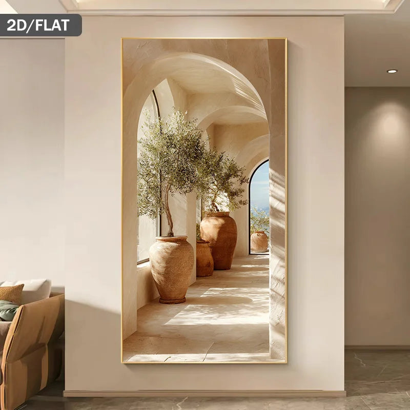 A large vertical framed wall art print hangs on a beige wall in a modern interior. The artwork shows a sunlit architectural corridor with arches, large terracotta-style pots, and olive trees.