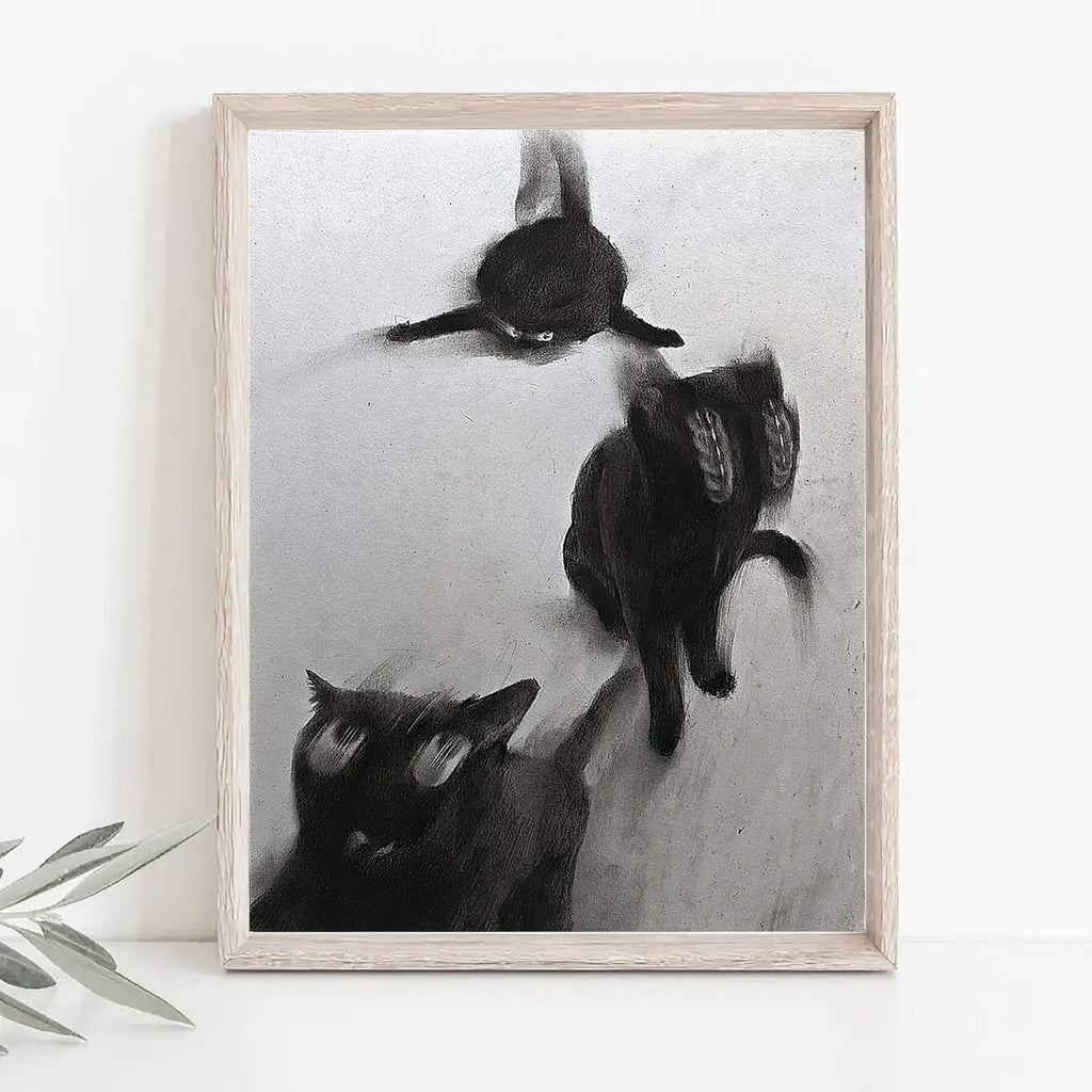 Black and white art print depicting three cats in motion, displayed on a white surface against a white wall with a small plant leaf visible at the bottom left.