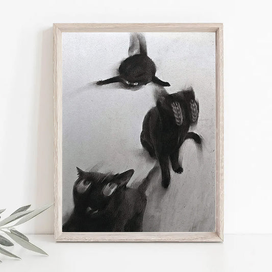 Black and white art print depicting three cats in motion, displayed on a white surface against a white wall with a small plant leaf visible at the bottom left.