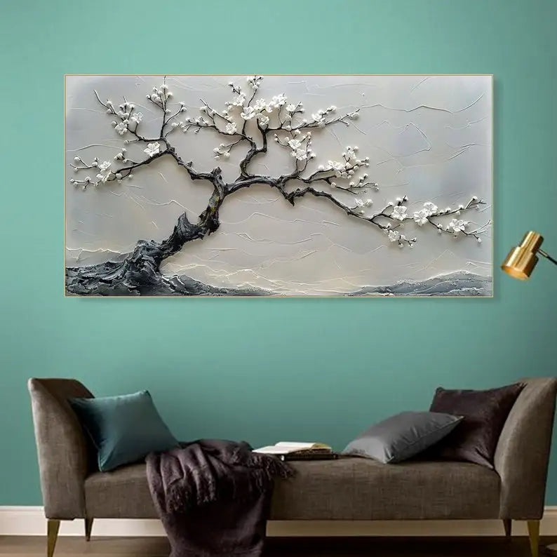 A large rectangular wall art piece featuring a stylized tree with white blossoms and dark textured branches, mounted on a teal wall above a gray sofa with pillows and a throw blanket.