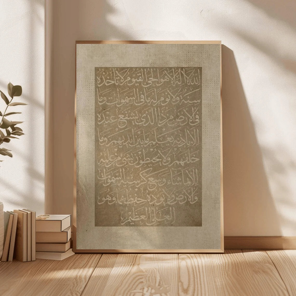 Framed artwork with Arabic calligraphy on a wooden floor with books and a plant.