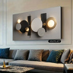 A modern abstract wall art piece mounted above a sofa in a living room. The artwork features overlapping circles, rectangles, and illuminated light bulbs in black, white, beige, and gold tones.