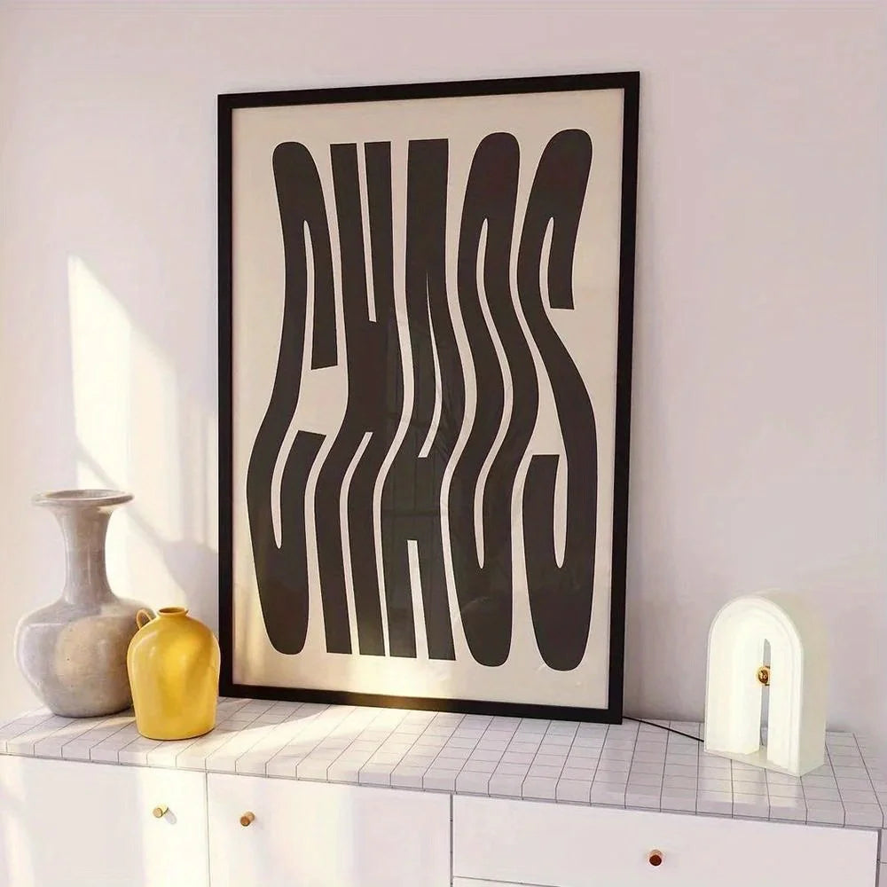 A framed abstract typographic wall art print with large black wavy lettering on a cream background, leaning on a white cabinet against a pale wall. Decorative vases and a small lamp sit beside it in a styled interior setting.