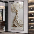 A large vertical framed wall art piece hangs on a light gray wall in a modern interior. The artwork depicts a stylized tree with white and beige branches and trunk, accented with small gold leaf-like details.