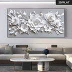 A large rectangular wall art piece showing a 3D-effect white floral design with layered roses, peonies, and leaves on a light gray background. The artwork is displayed above a modern beige sofa in a living room setting.