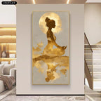 A vertical framed wall art print showing a meditating woman silhouette in gold tones against a pale background, displayed in a modern interior hallway with a staircase and sofa visible.