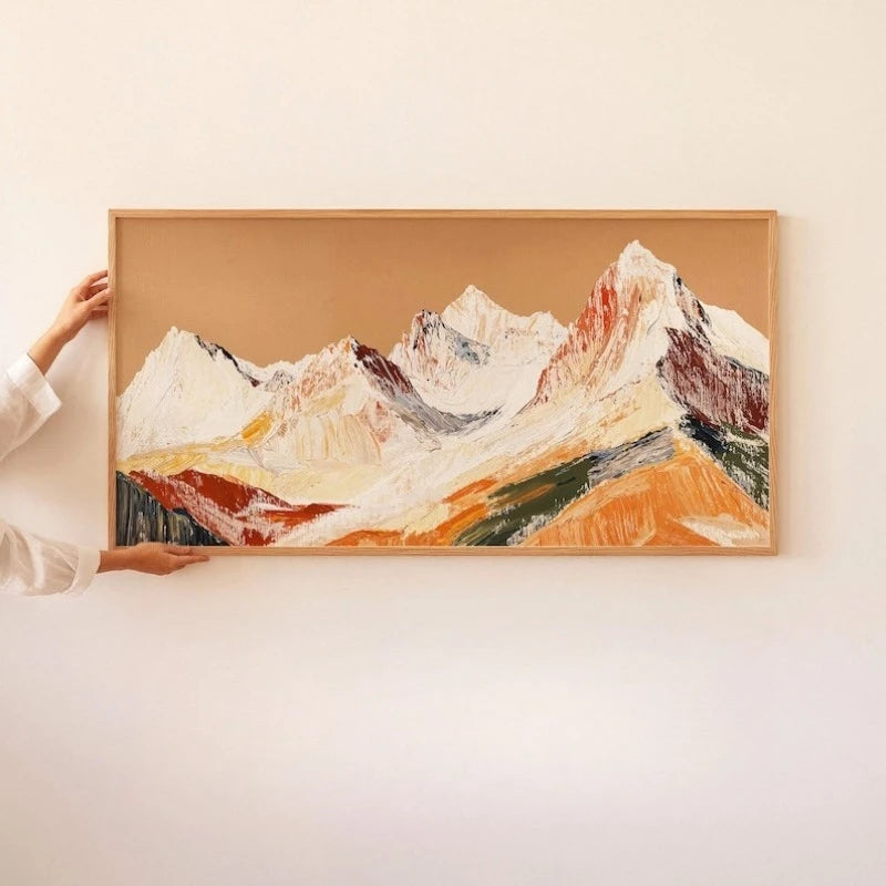 A landscape painting of snow-capped mountains with warm ochre, white, and green brushstrokes, held by hands against a plain off-white wall.