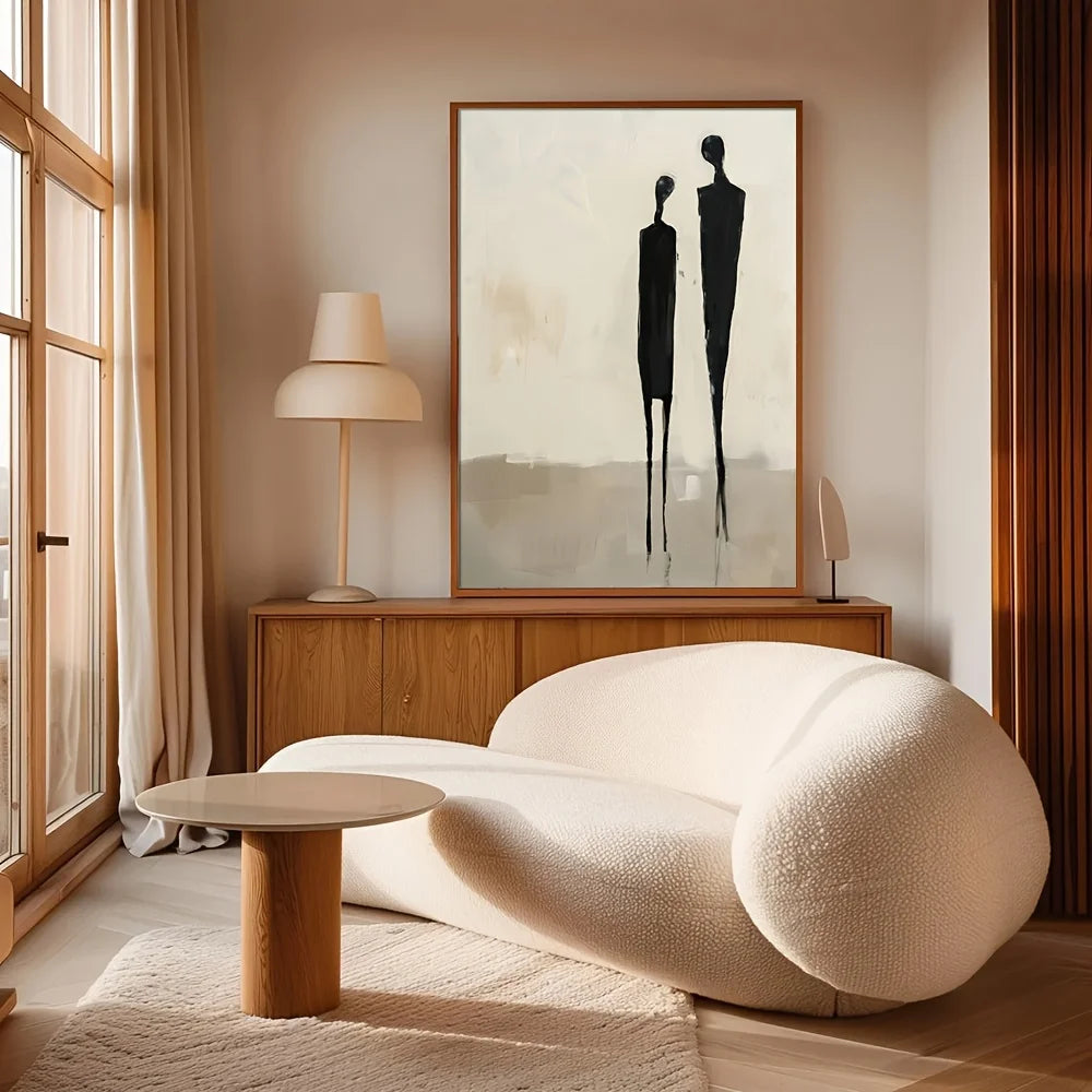 A modern living room scene featuring a large framed abstract art print with two black minimalist human figures on a warm off-white background, hung above a wooden sideboard with a cream-colored curved sofa and a small round wooden table in the foreground.