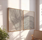 Two abstract wall art pieces with wavy lines on a light wall, with a plant and chair in the foreground.