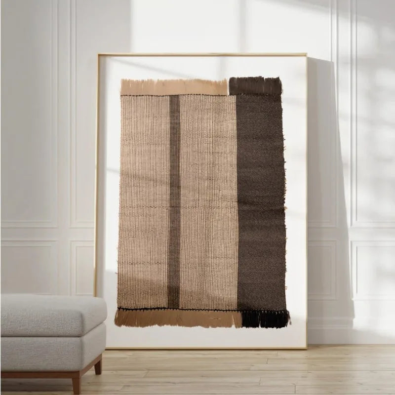 A large framed abstract textile artwork leaning against a white paneled wall in a bright room. The piece features vertical woven bands in beige, tan, brown, and dark brown with rough fringe at the top and bottom.