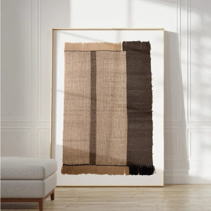 A large framed abstract textile artwork leaning against a white paneled wall in a bright room. The piece features vertical woven bands in beige, tan, brown, and dark brown with rough fringe at the top and bottom.