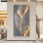 A vertical framed wall art piece featuring large white feathers with warm gold highlights, displayed in a modern interior hallway with a staircase and built-in shelving visible.