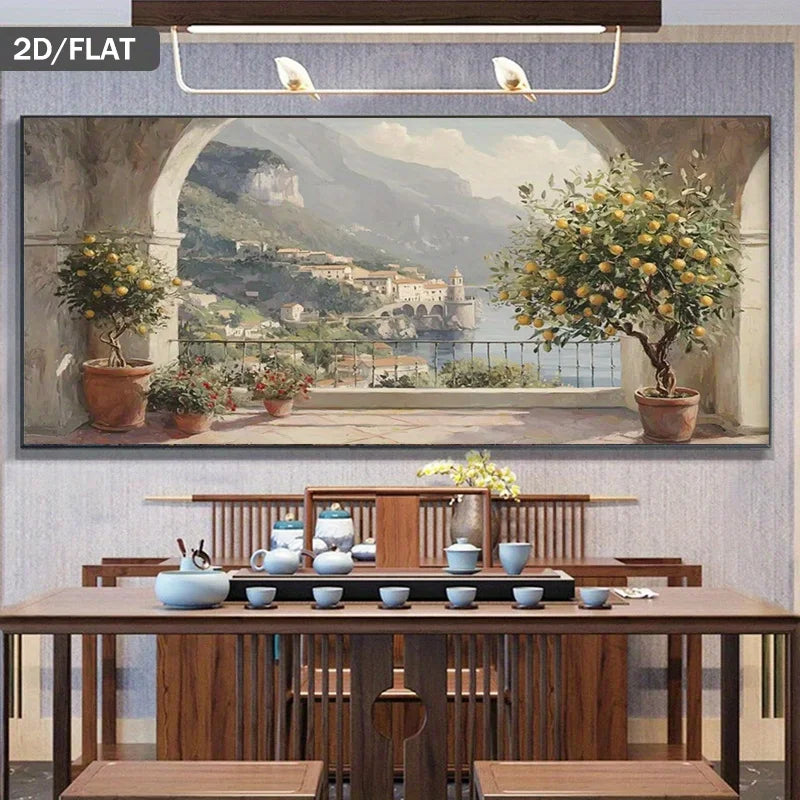 A large framed wall art print depicting a scenic coastal town with mountains, water, and lemon trees, displayed above a wooden dining table in a styled interior.