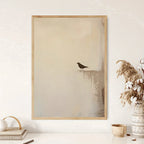 Minimalist art print featuring a small black bird perched on a ledge against a soft beige and gray background, displayed on a white wall above a styled tabletop with books, a candle, and dried grass in a vase.