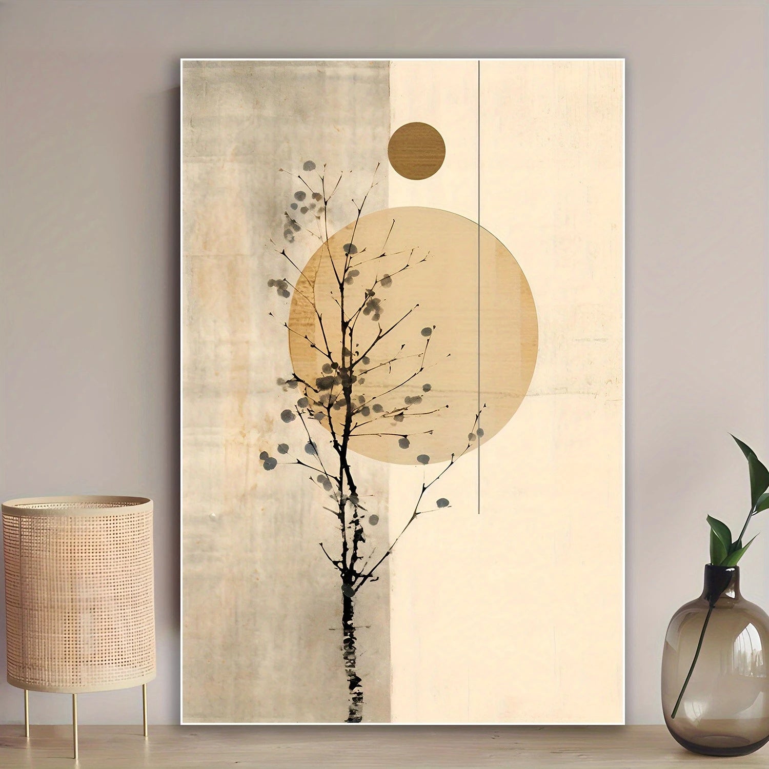 A vertical framed wall art print featuring an abstract black tree silhouette over large beige and cream circles, displayed on a tabletop against a light gray wall with a woven lamp on the left and a glass vase with green leaves on the right.