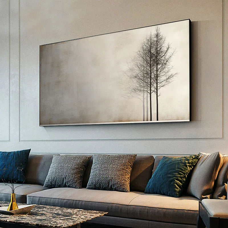 A large rectangular wall art print featuring three bare trees against a misty beige background, displayed above a gray sofa with blue and gray pillows in a modern living room setting.