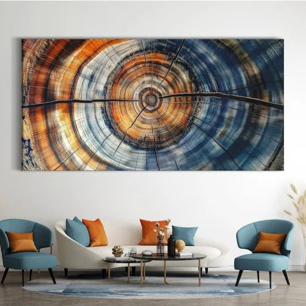 A large rectangular wall art piece featuring a circular wood grain or tree-ring style abstract design in blue, orange, brown, and cream tones, displayed above a modern white sofa in a styled living room with blue chairs and decorative plants.