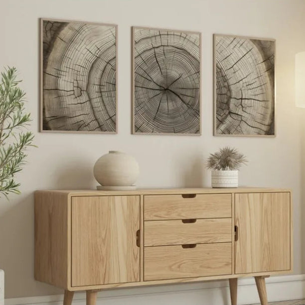 A mid-century modern light wood sideboard cabinet styled in a living room setting, featuring three framed wood grain wall art prints above it, a ceramic vase, and a small potted plant.