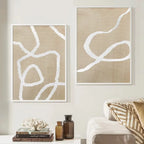 Two abstract canvas prints with white organic line art on a beige linen-textured background, displayed on a light wall above a sofa with decorative pillows and small bottles.