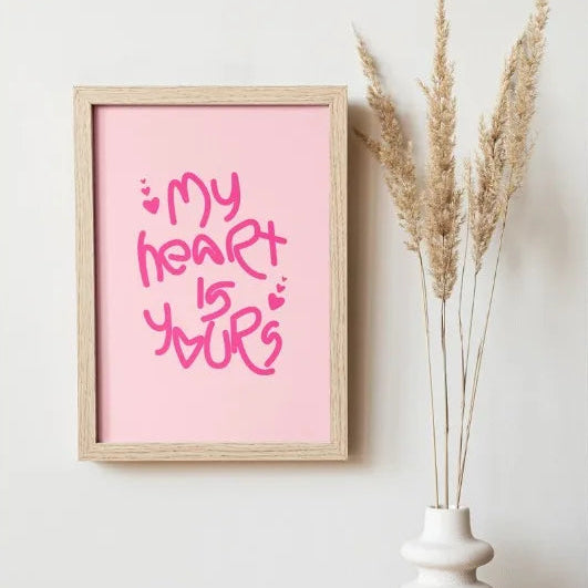 A framed pink art print with the phrase 'my heart is yours' in pink script, displayed on a light wall above a wooden surface next to a white vase with dried pampas grass.