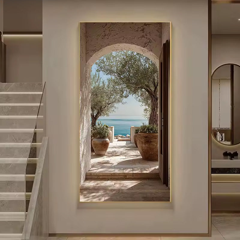 A vertical wall art print depicting a Mediterranean courtyard with an olive tree and sea view, mounted on a beige wall in a modern interior setting.
