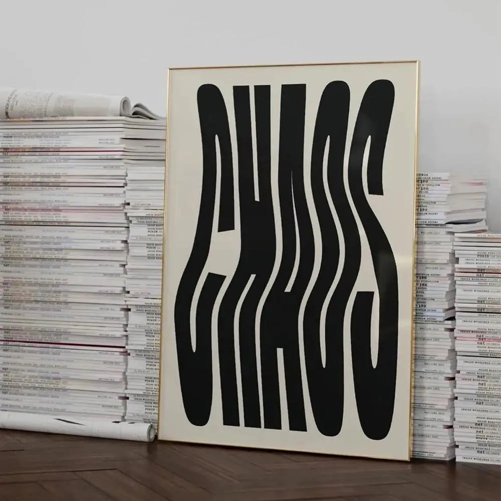 A framed abstract art print with bold black wavy lettering on a cream background, leaning against stacks of magazines on a wooden floor.
