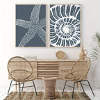 Two framed wall art prints hang above a light wood console in a styled interior. The left print shows a white starfish on a dark blue-gray background, and the right print shows a white spiral shell pattern on the same blue-gray background.