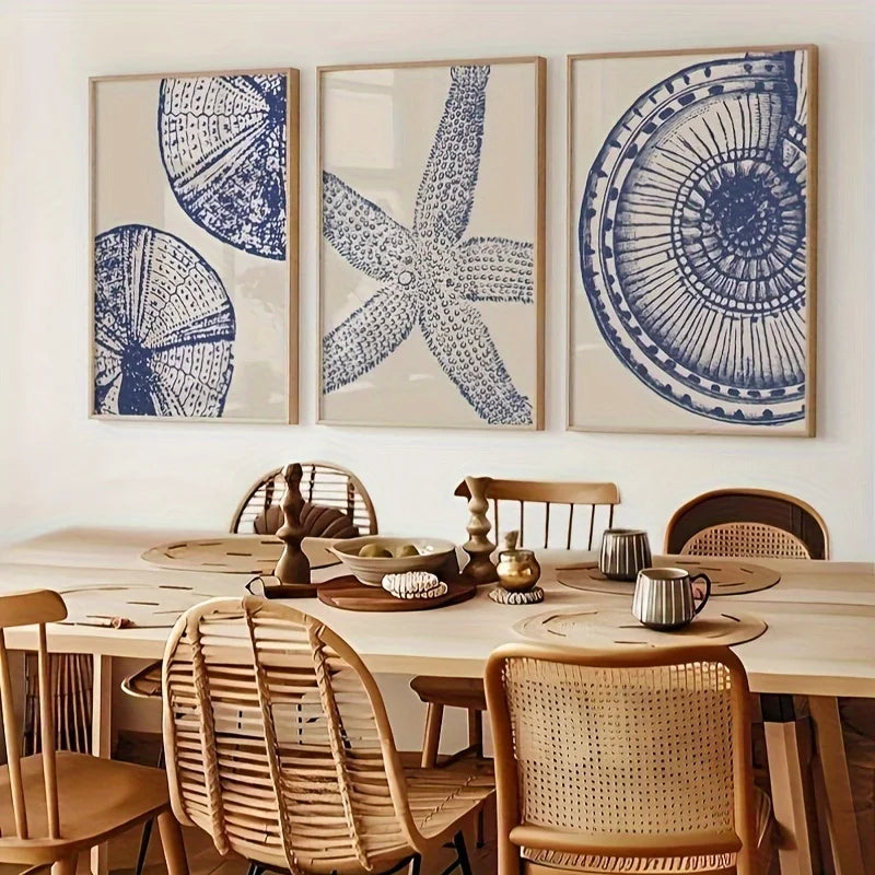 Three framed vertical art prints with blue nautical sketches of a starfish and seashells on a beige background, displayed on a white wall above a wooden dining table with woven chairs.