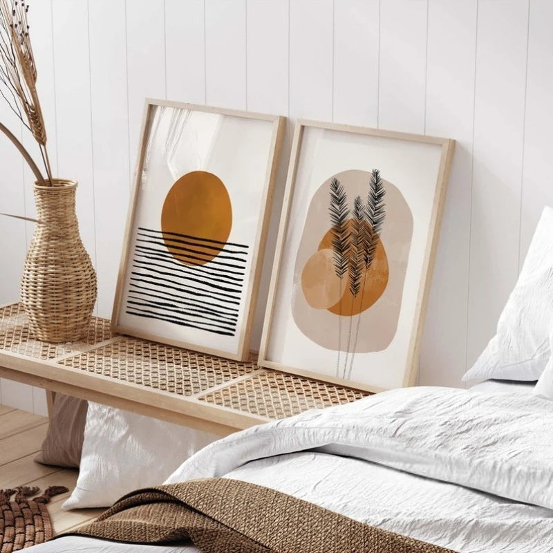 Two framed abstract botanical art prints leaning on a woven bench beside a bed with white bedding and a wicker vase with dried stems.