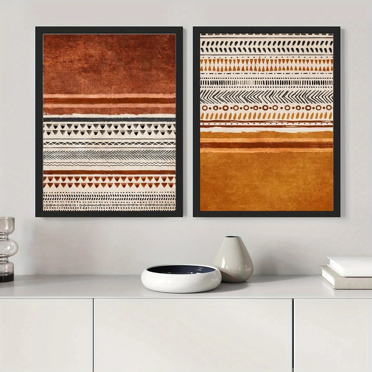 Two framed vertical wall art prints with horizontal geometric and textured patterns in rust, terracotta, cream, and orange tones, displayed side by side on a light gray wall above a white console table with decorative objects.