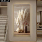 A framed wall art print displayed in a modern interior hallway. The artwork shows three beige and brown ceramic vases filled with tall cream pampas grass and small dried red stems, set against a light wood plank background with a thin gold frame.