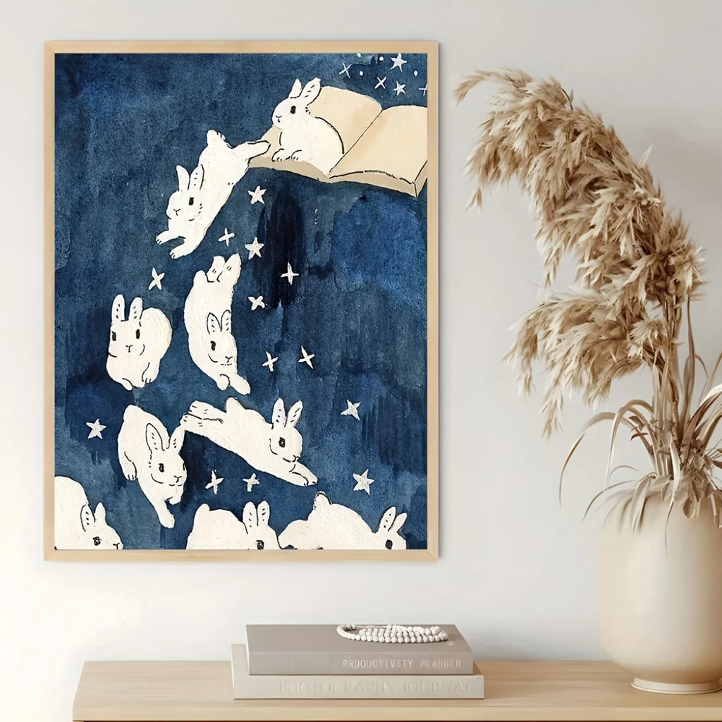Wall art print featuring white rabbits floating in a starry navy blue sky, hanging on a light wall above a wooden shelf with books and a vase of dried grass.