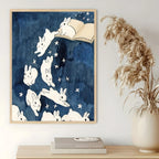 Wall art print featuring white rabbits floating in a starry navy blue sky, hanging on a light wall above a wooden shelf with books and a vase of dried grass.