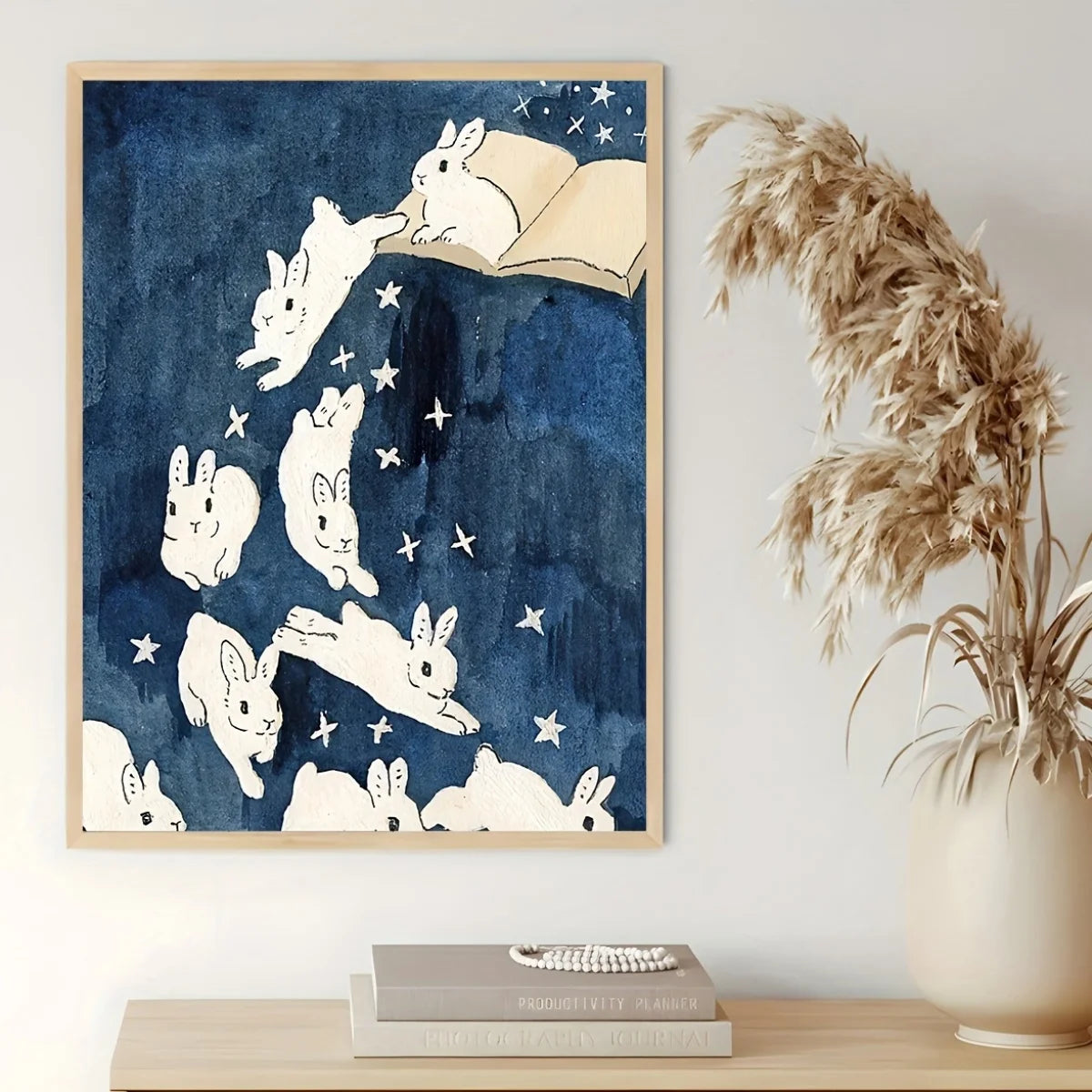 Wall art print featuring white rabbits floating in a starry navy blue sky, hanging on a light wall above a wooden shelf with books and a vase of dried grass.