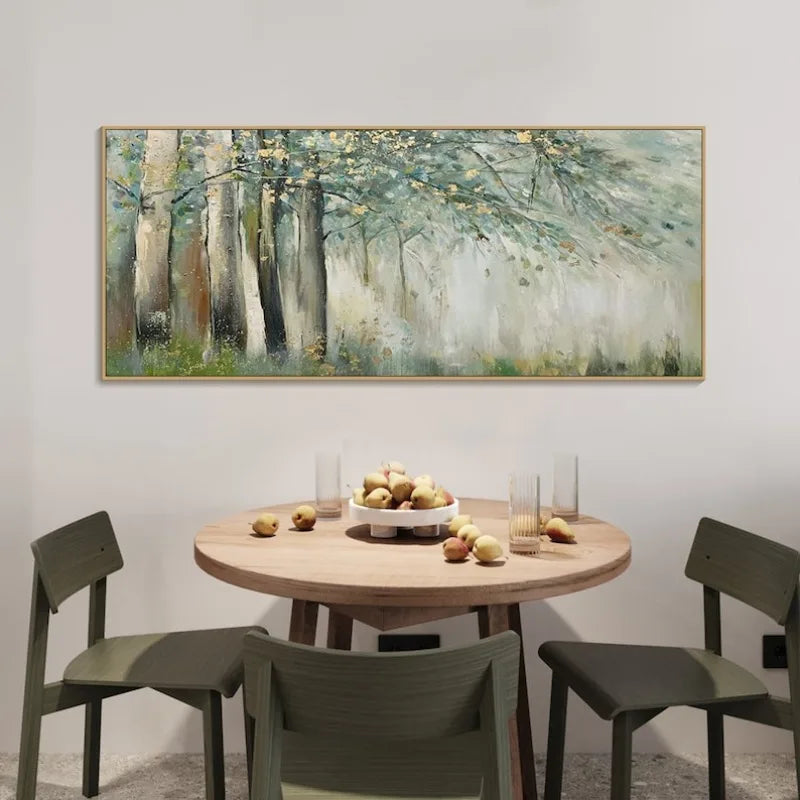 A large horizontal framed painting of a misty forest with tall trees hangs on a light gray wall above a round wooden dining table with four chairs and a bowl of fruit.